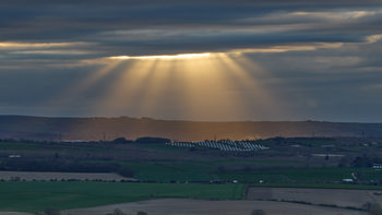 This landscape photograph captures striking Gods rays, also known as crepuscular rays, shining through heavy clouds on a spring evening over the rural countryside near Bolsover in Derbyshire, United Kingdom. The scene is dominated by the dramatic beams of sunlight breaking through the cloud cover, illuminating a solar farm with rows of solar panels in the middle distance. The photo encapsulates the contrast between the natural beauty of the rolling English fields and the modern presence of renewable energy infrastructure in rural England. The combination of evening light, extensive cloud formations, and visible solar panels makes this a characteristic view of the Derbyshire countryside.